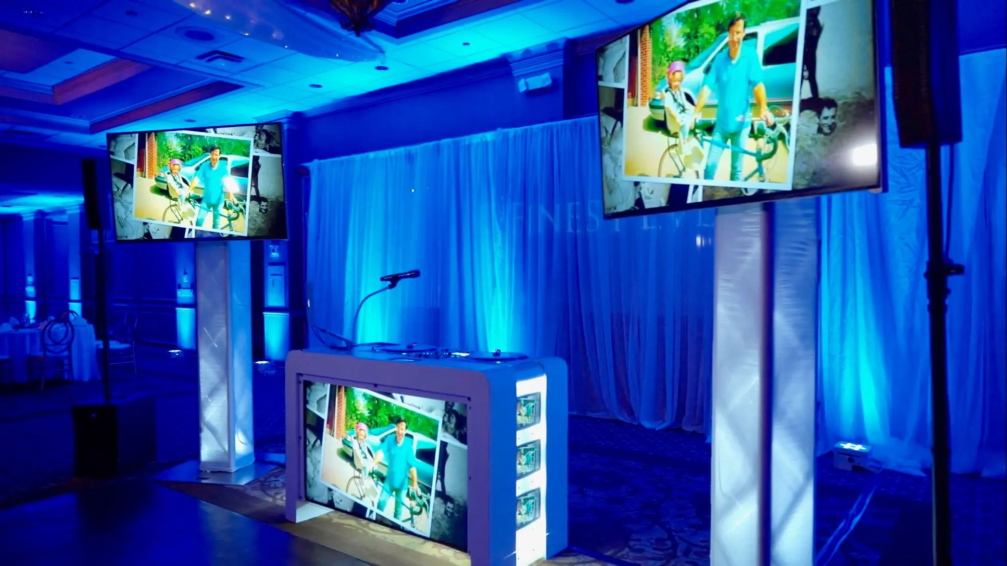 Video DJ booth and truss-mounted TVs showing wedding photos under blue uplighting